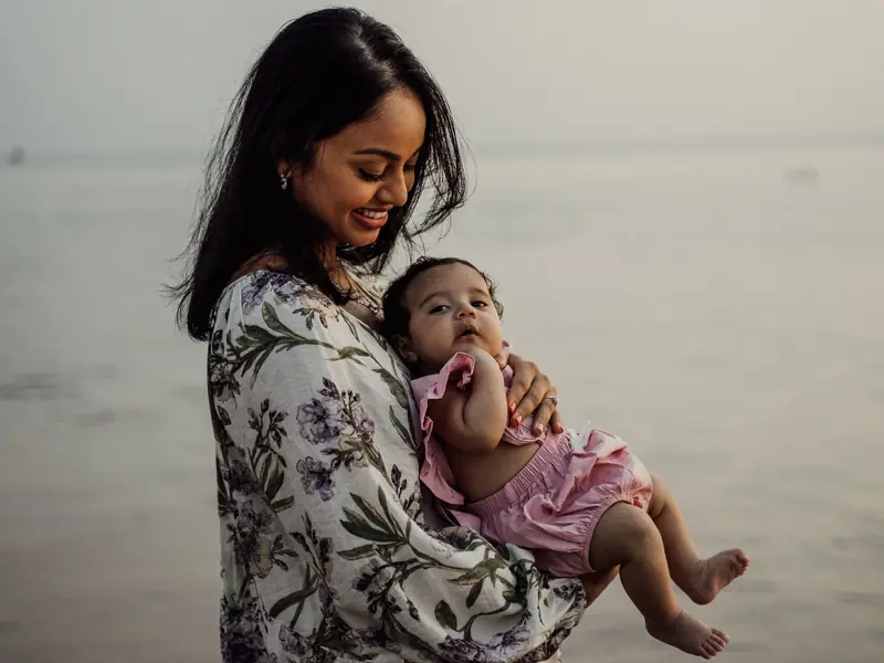 A smiling mother holds her baby in her arms by the water in soft evening light