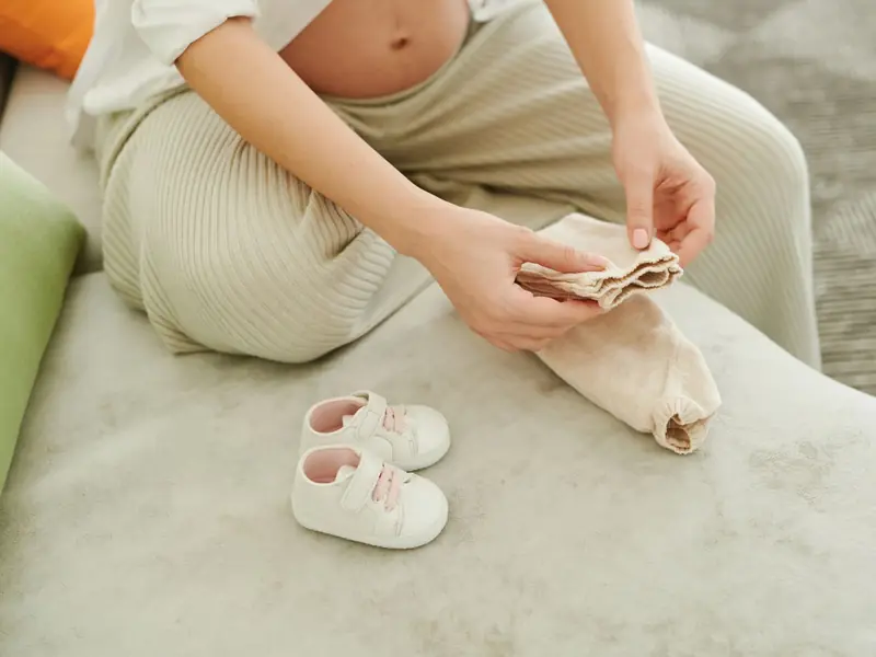 A pregnant woman folds tiny baby clothes next to a pair of baby shoes