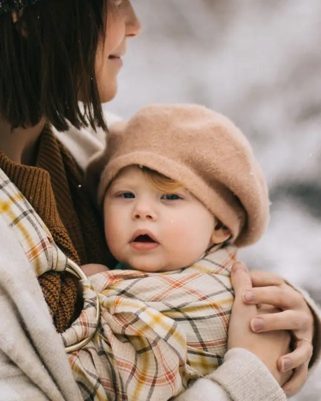 A baby in a beret rides in a ring sling, face clear and close